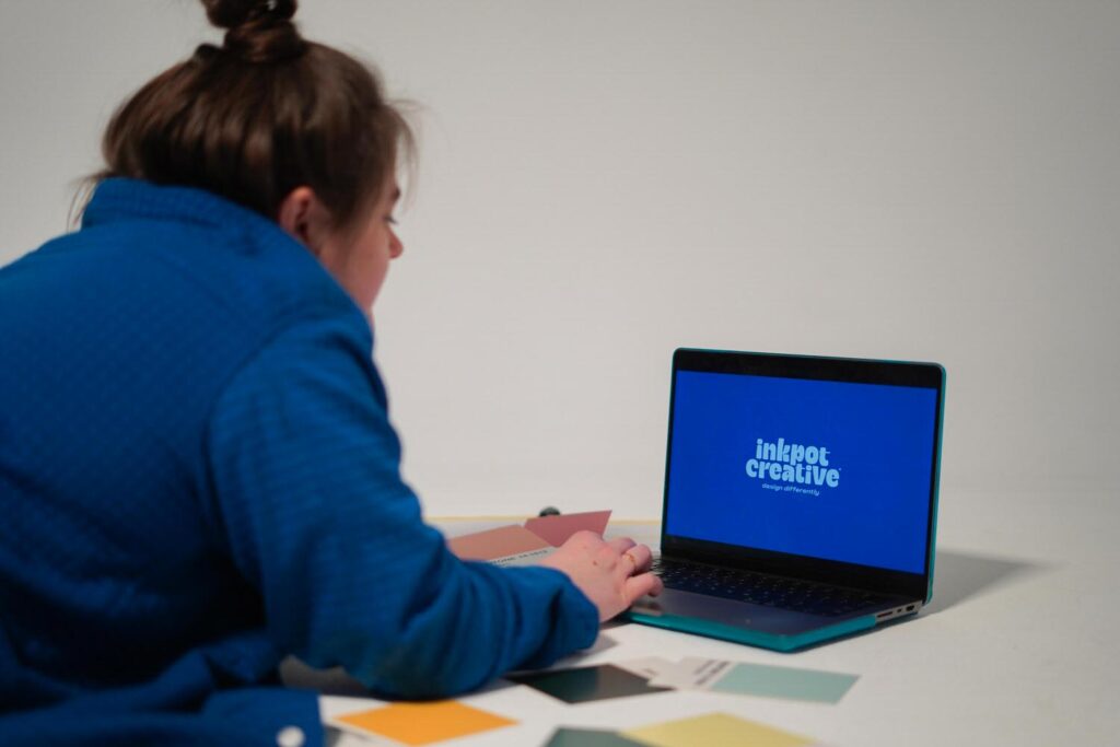 A person in a blue shirt, viewed from the back, working on a laptop that displays the text "inkpot creative" on the screen, surrounded by various Pantone color cards.