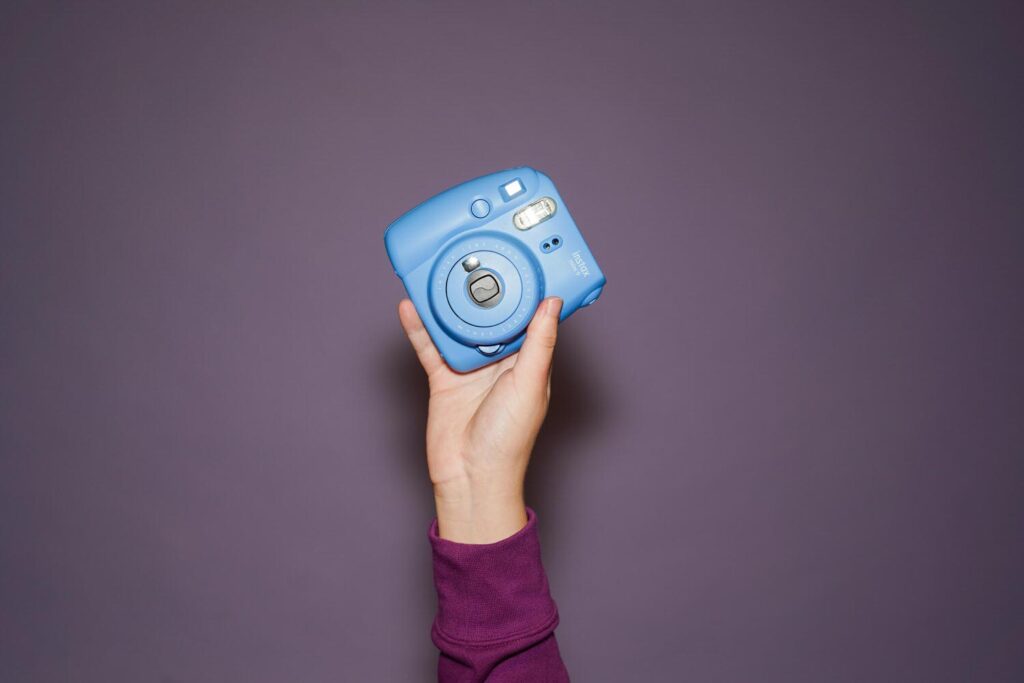 A hand holding a blue Instax Mini 9 instant camera against a plain purple background.