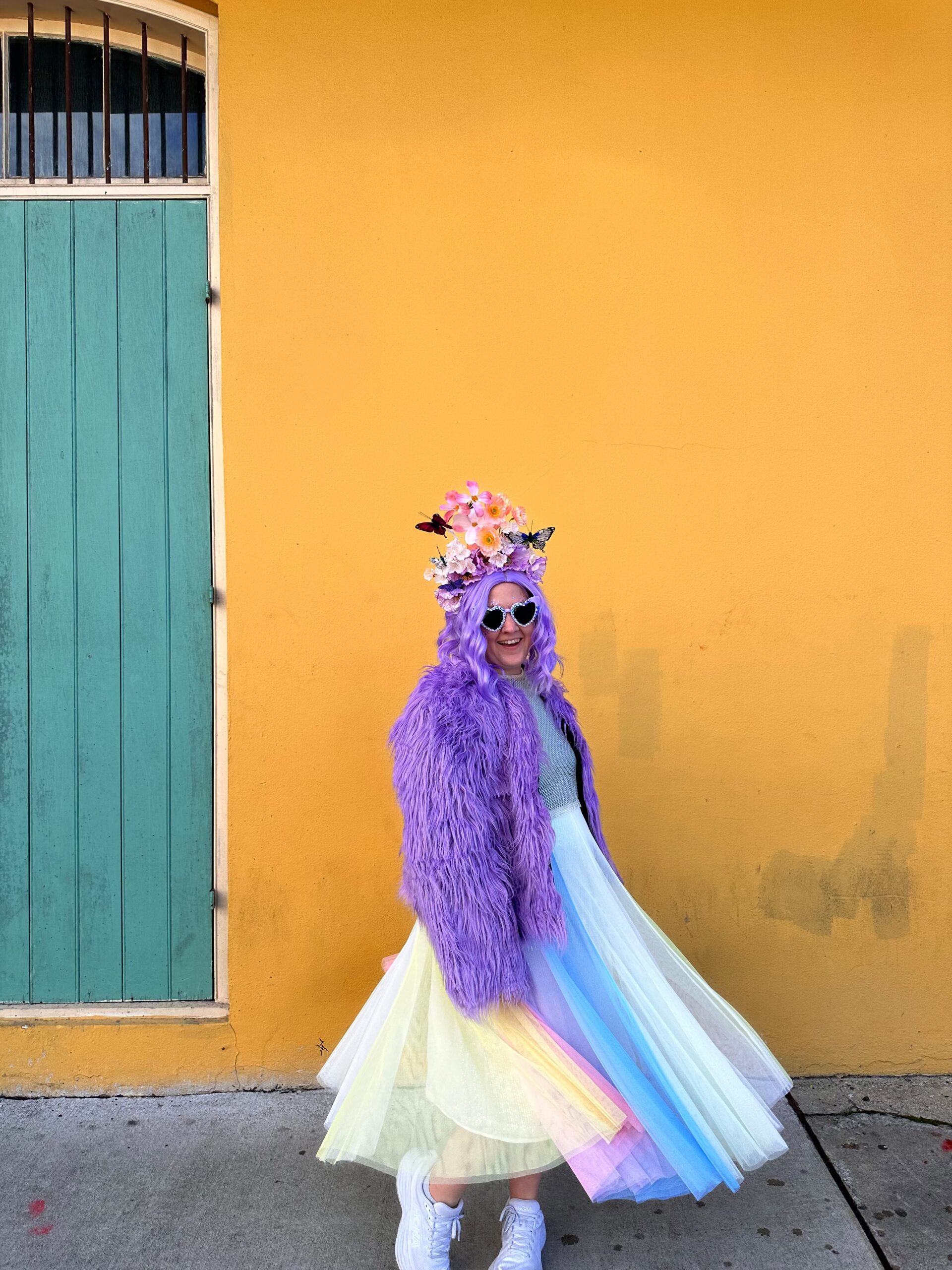 A person in a rainbow dress and purple jacket standing in front of a colorful wall