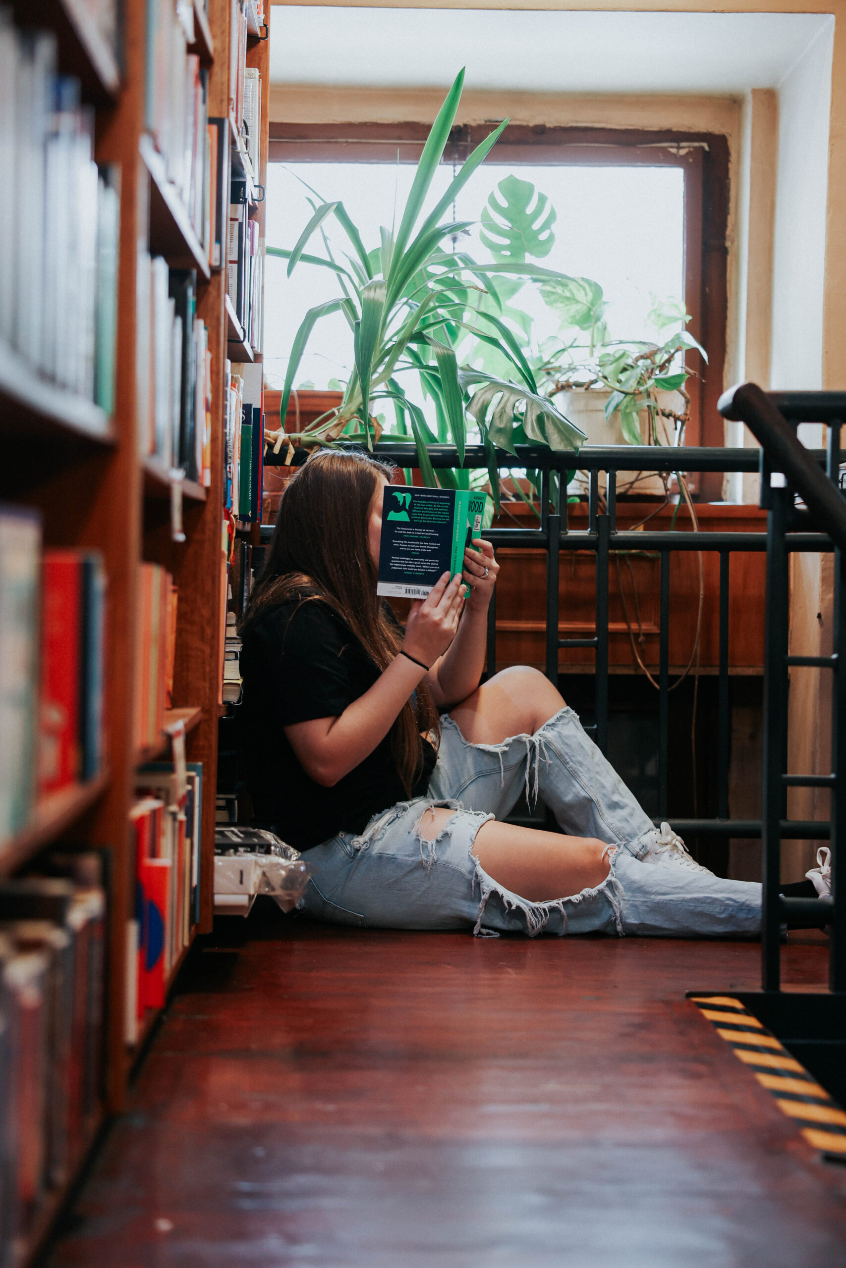 a woman wearing jeans and a t-shirt holding a book and sitting in the corner of a bookstore reading it 