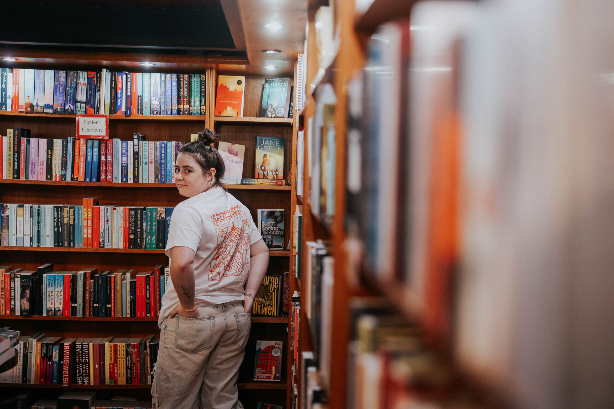a woman with her hands in her pockets looking over her shoulder while standing in a bookstore 