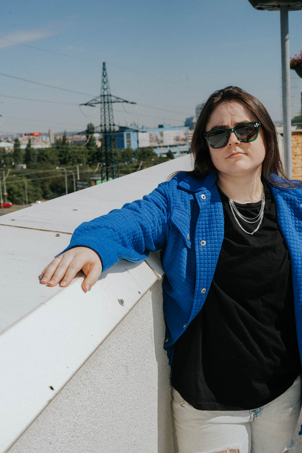 a woman wearing a black shirt, chain necklace, black sunglasses and blue jacket posing with her arm against a wall