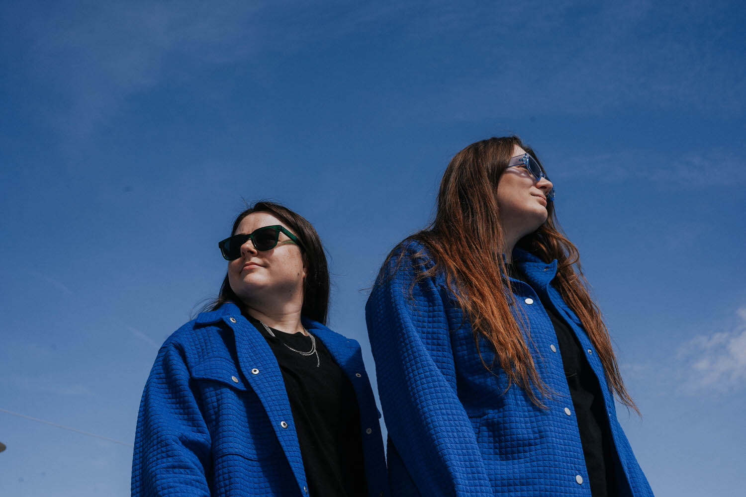 two women wearing blue jackets and sunglasses standing next to each other with a blue sky behind them