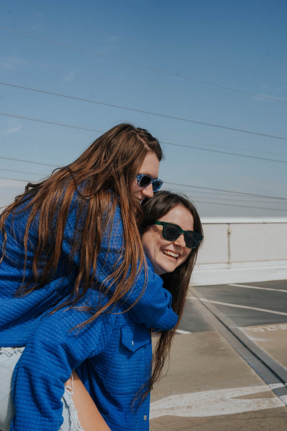 two women on top of a rooftop of a parking garage both wearing matching blue jackets one woman is piggybacking on the other woman's back and they are both laughing