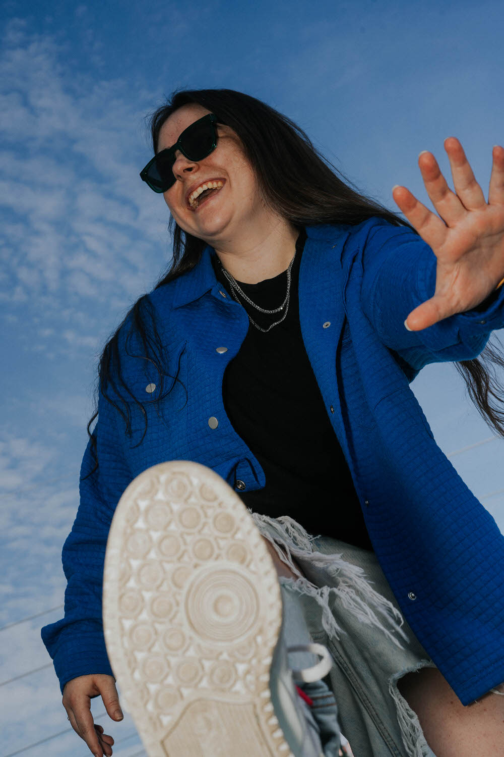 a woman laughing wearing a blue jacket, a chain necklace, black shirt and sunglasses with a blue sky behind her