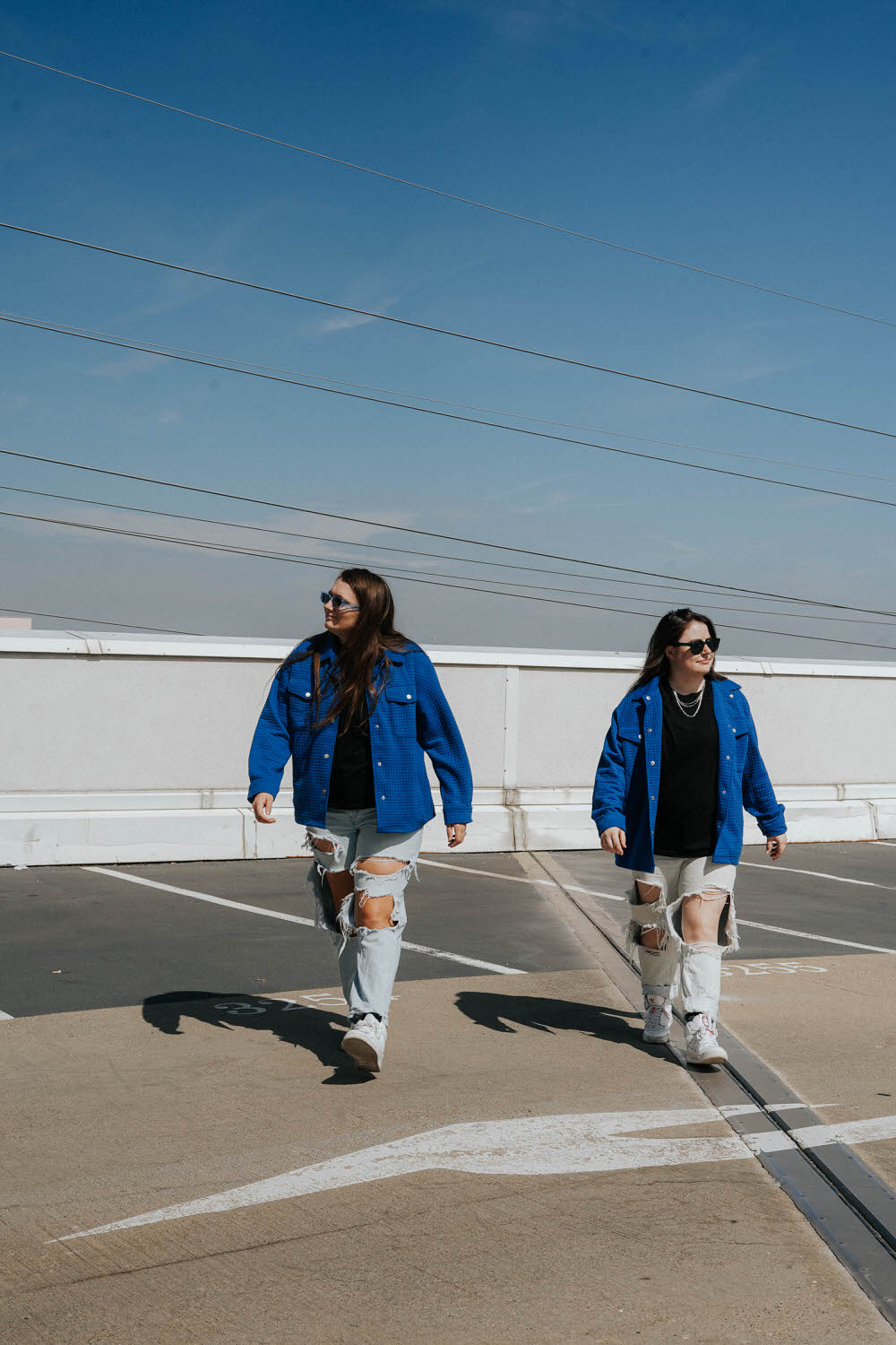 two women walking side by side wearing matching outfits with ripped jeans, black shirts and blue jackets and sunglasses they are on the rooftop of a parking garage