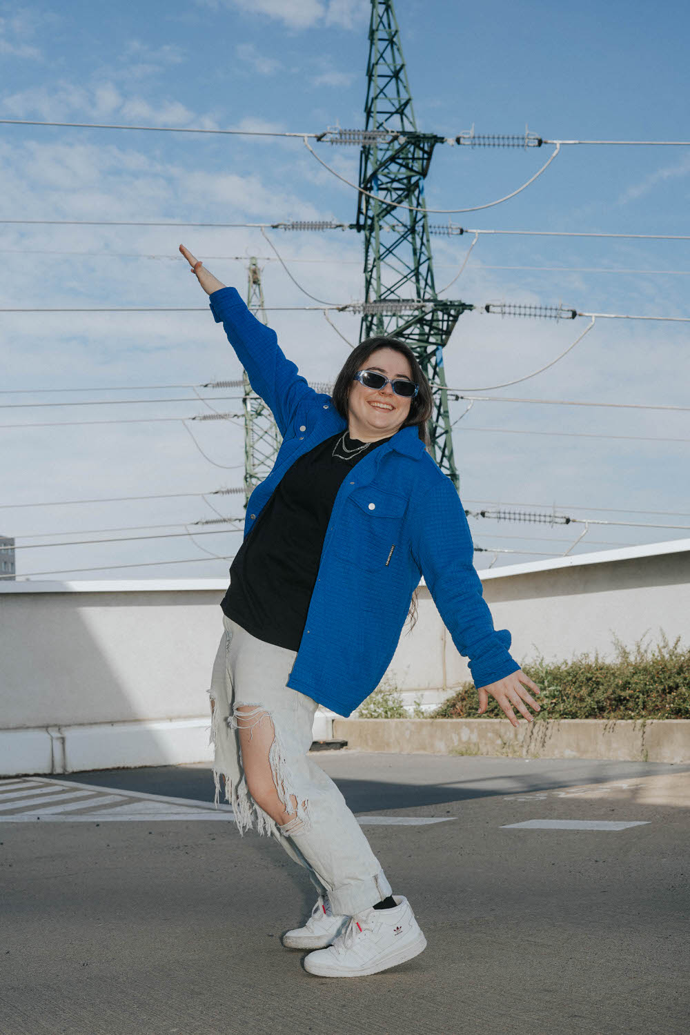 a woman posing playfully on the rooftop of a parking garage wearing a blue jacket, black shirt, sunglasses, and ripped jeans