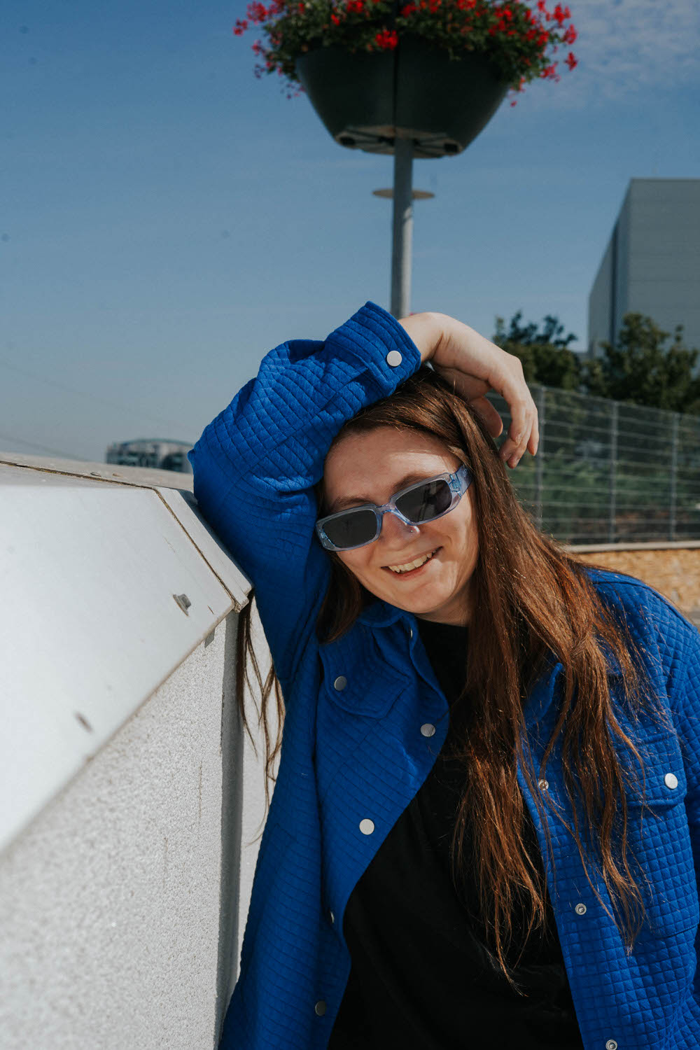 a woman on the rooftop of a parking garage posing next to the wall with her hand above her head wearing blue sunglasses and a blue jacket