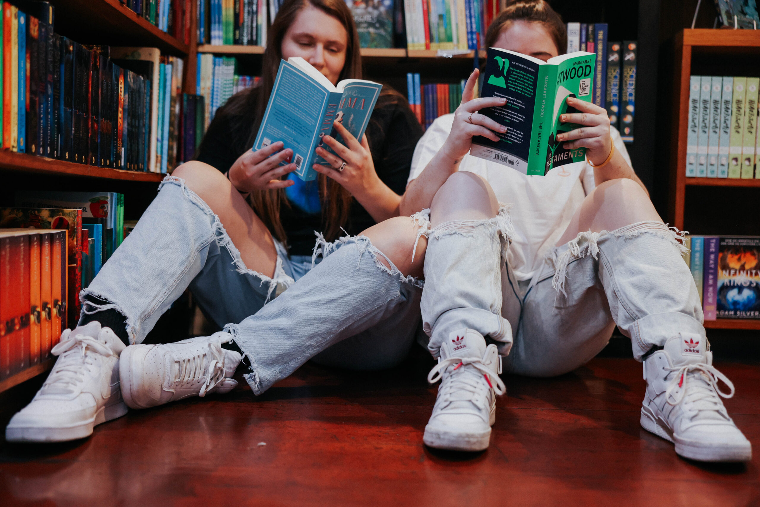 two people sitting side by side on the floor of a bookstore in front of a bookshelf reading books 